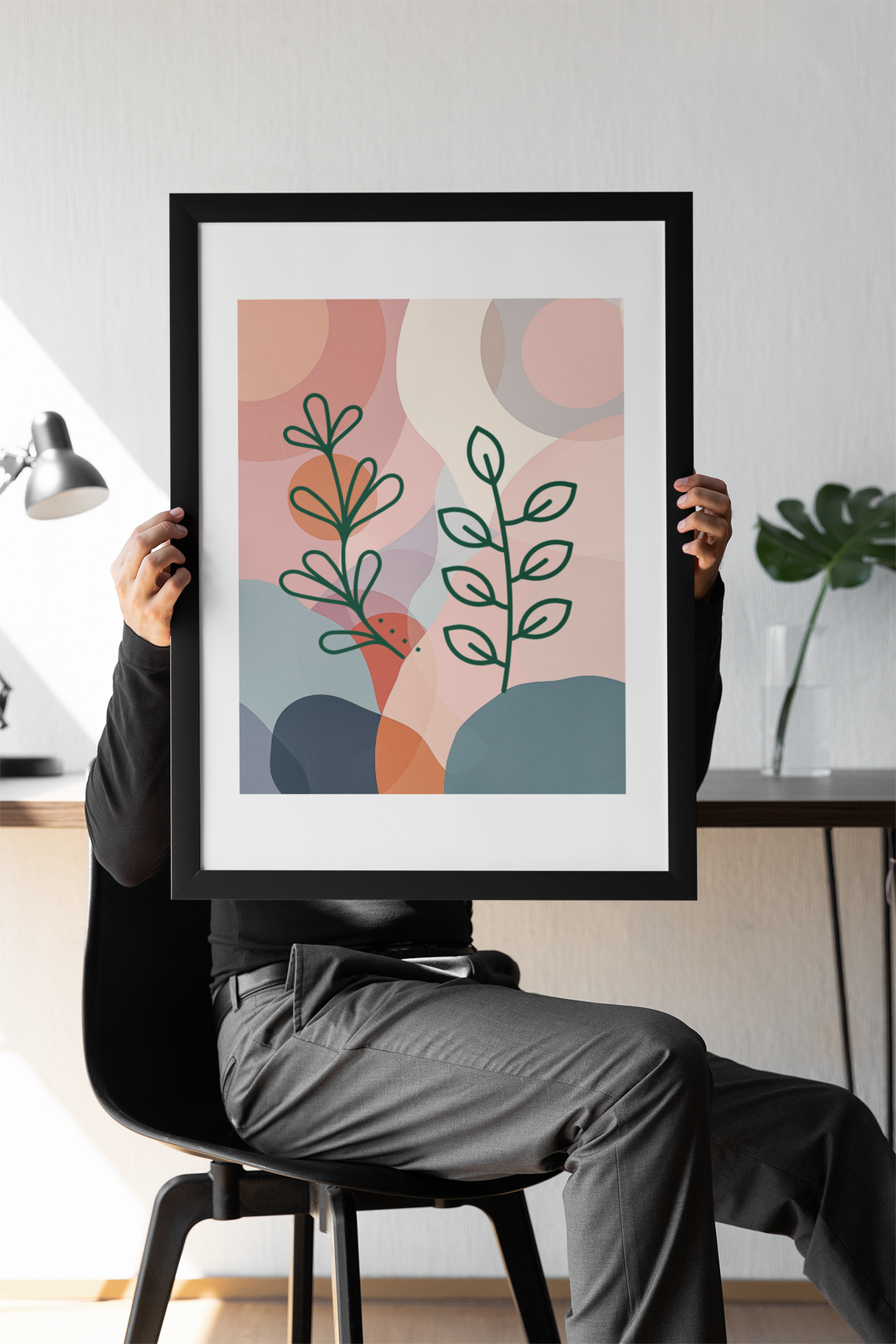 Person holding a framed abstract art print with plants in a minimalistic room.
