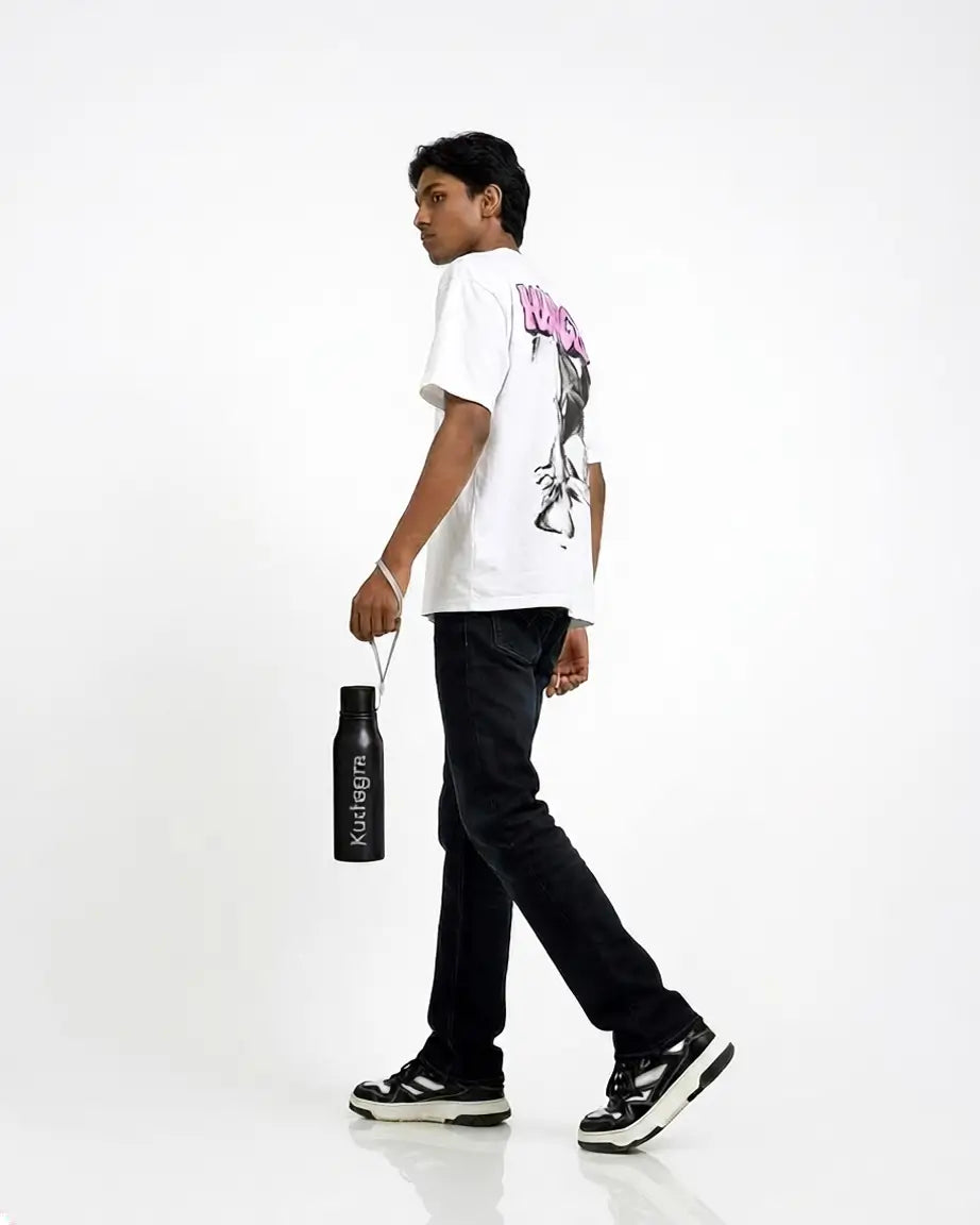 A young man walking in profile wearing a white graphic t-shirt, black jeans, and black-and-white sneakers, holding a black water bottle with the brand name &
