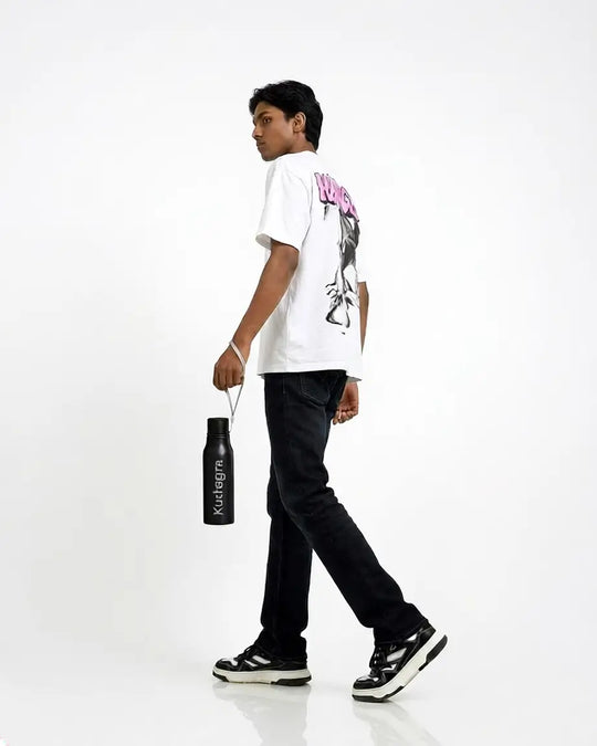 A young man walking in profile wearing a white graphic t-shirt, black jeans, and black-and-white sneakers, holding a black water bottle with the brand name &