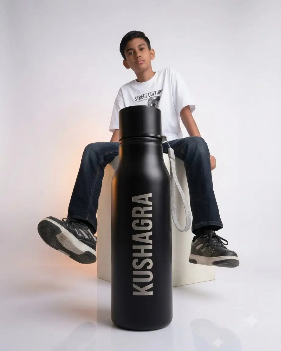 A boy wearing a white graphic t-shirt and dark jeans sits on a white block, with a large matte black Kushagra insulated water bottle placed in the foreground against a clean white studio background.