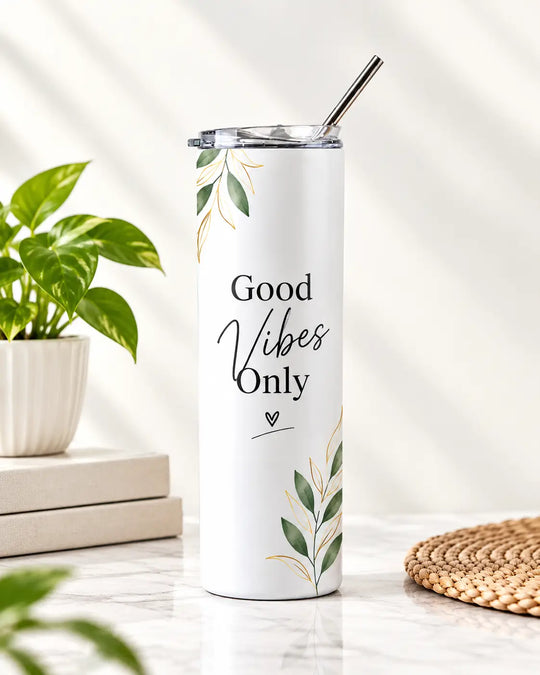 A tall white insulated tumbler with a metal straw stands on a light marble surface. The tumbler has green leaf illustrations and the phrase &