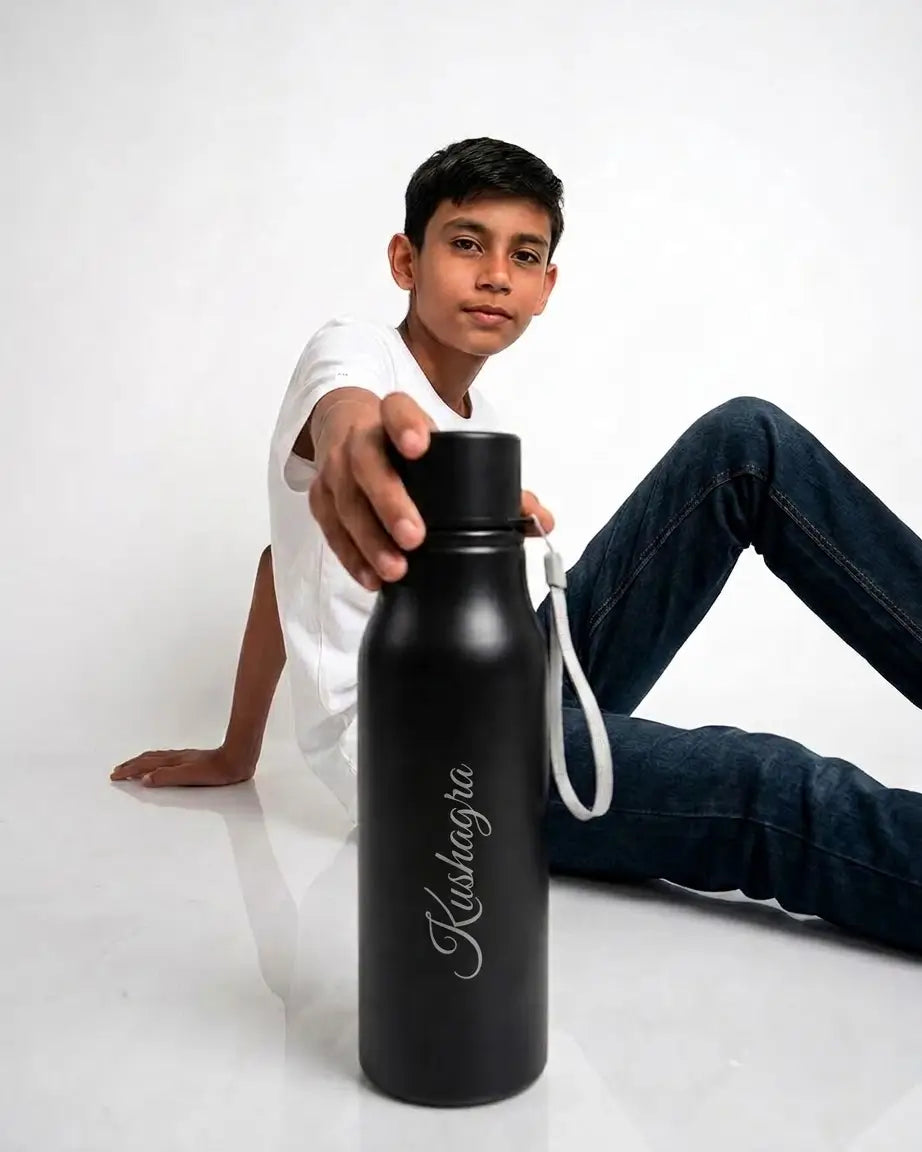 A boy in a white t-shirt and dark blue jeans sits on a white floor, holding a matte black insulated water bottle with a white strap and the brand name 'Kulhagra' printed on it.