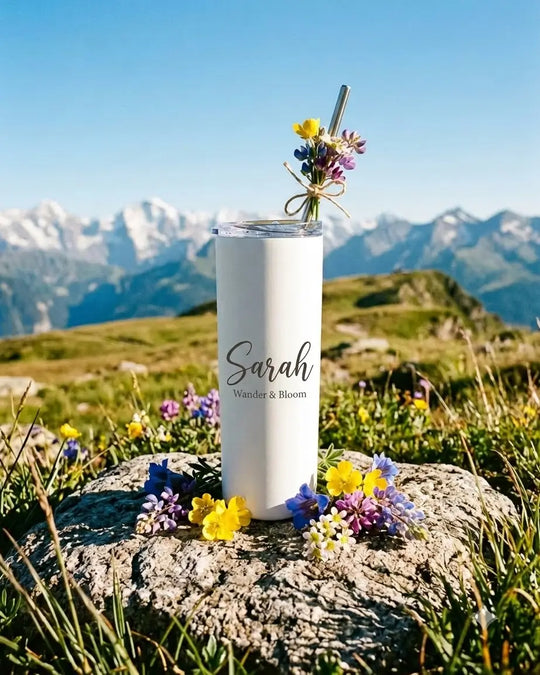 Custom name engraved white skinny tumbler with lid and steel straw, placed on a rock among alpine wildflowers with snow-capped mountains behind