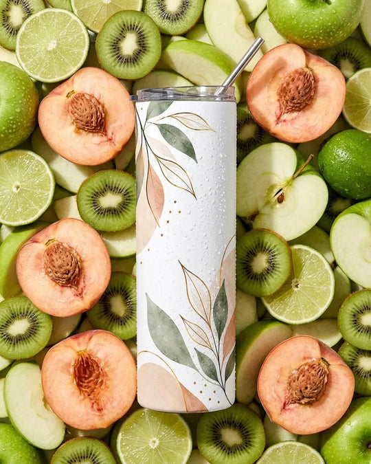 White skinny tumbler with clear lid and metal straw, printed with sage-green and blush-peach watercolor leaves and gold line accents, standing among sliced peaches, green apples, kiwis and limes with water droplets on the surface.
